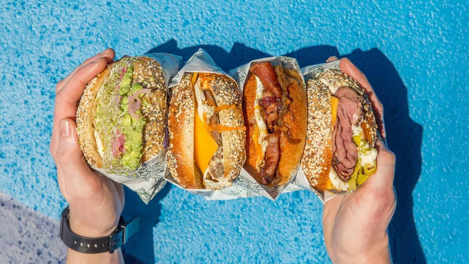 Two hands hold four different sandwiches with various fillings, including lettuce, cheese, bacon, and deli meats, against a bright blue background—perfect fuel for your Heart of Glass: A Weekend in Tacoma adventure.