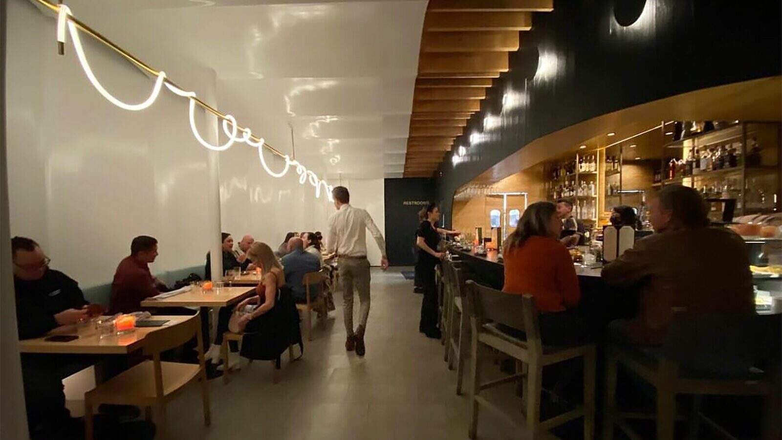 A restaurant interior with diners seated at tables and the bar, a person walking through the center aisle, and modern lighting along the wall—where As Smoke Fades, Hope Rises in L.A. is brought to life.