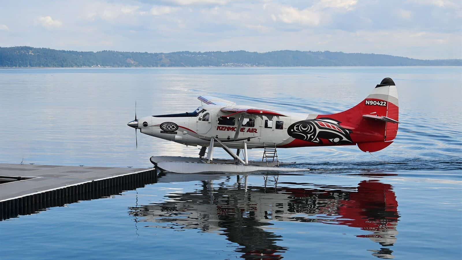 A red and white Kenmore Air floatplane is docked on calm water near a shoreline, with trees and hills in the background—capturing the serene spirit of Heart of Glass: A Weekend in Tacoma.