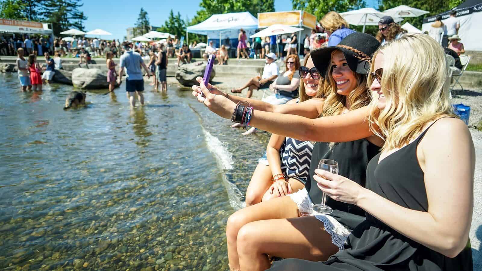 Three women sit by a lakeshore taking a selfie, surrounded by a crowd at an outdoor event with tents—set your sights on these summer festivals across Puget Sound.