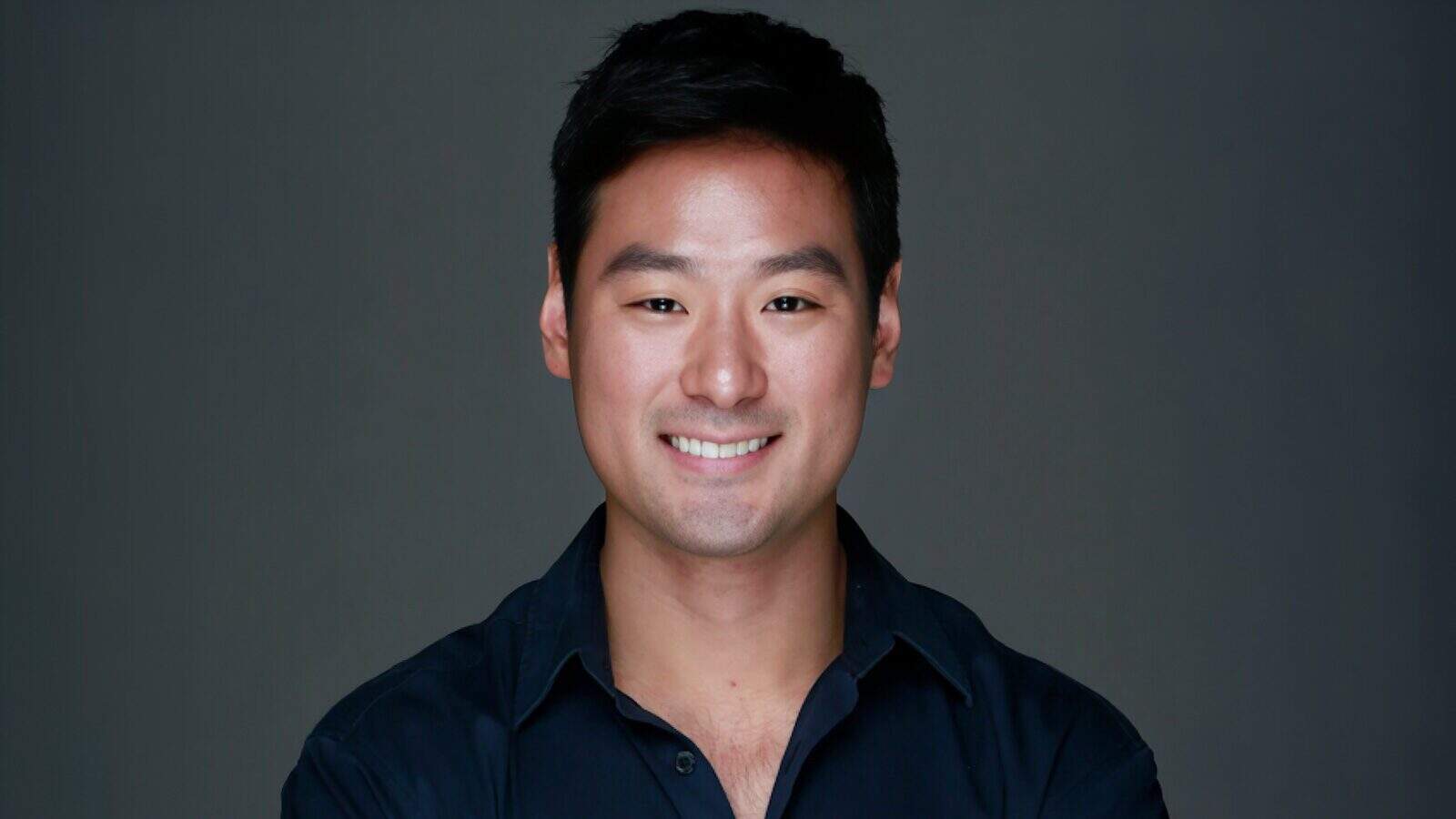 A man with short black hair and a dark shirt smiles at the camera against a plain dark background—Music Producer Kyu Lee talks Seattle, UW, and his Hollywood career.