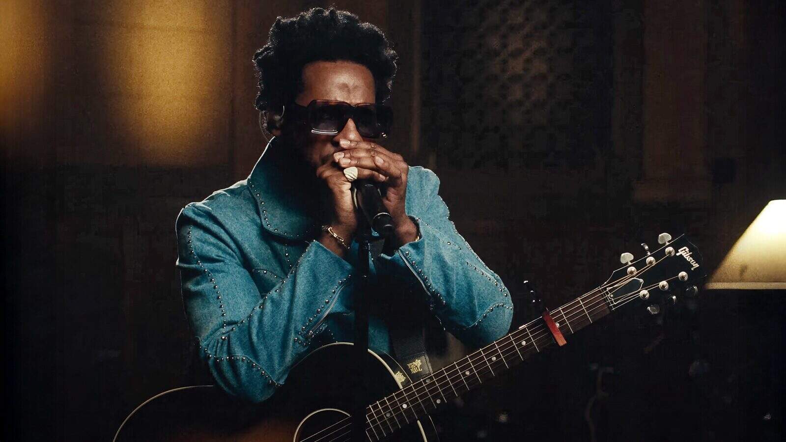 LeonBridges-ThatsWhatILove_1600x900 A musician wearing sunglasses and a blue jacket plays the harmonica and acoustic guitar on stage at Pioneer Square—perfect for those seeking unique things to do in Seattle.