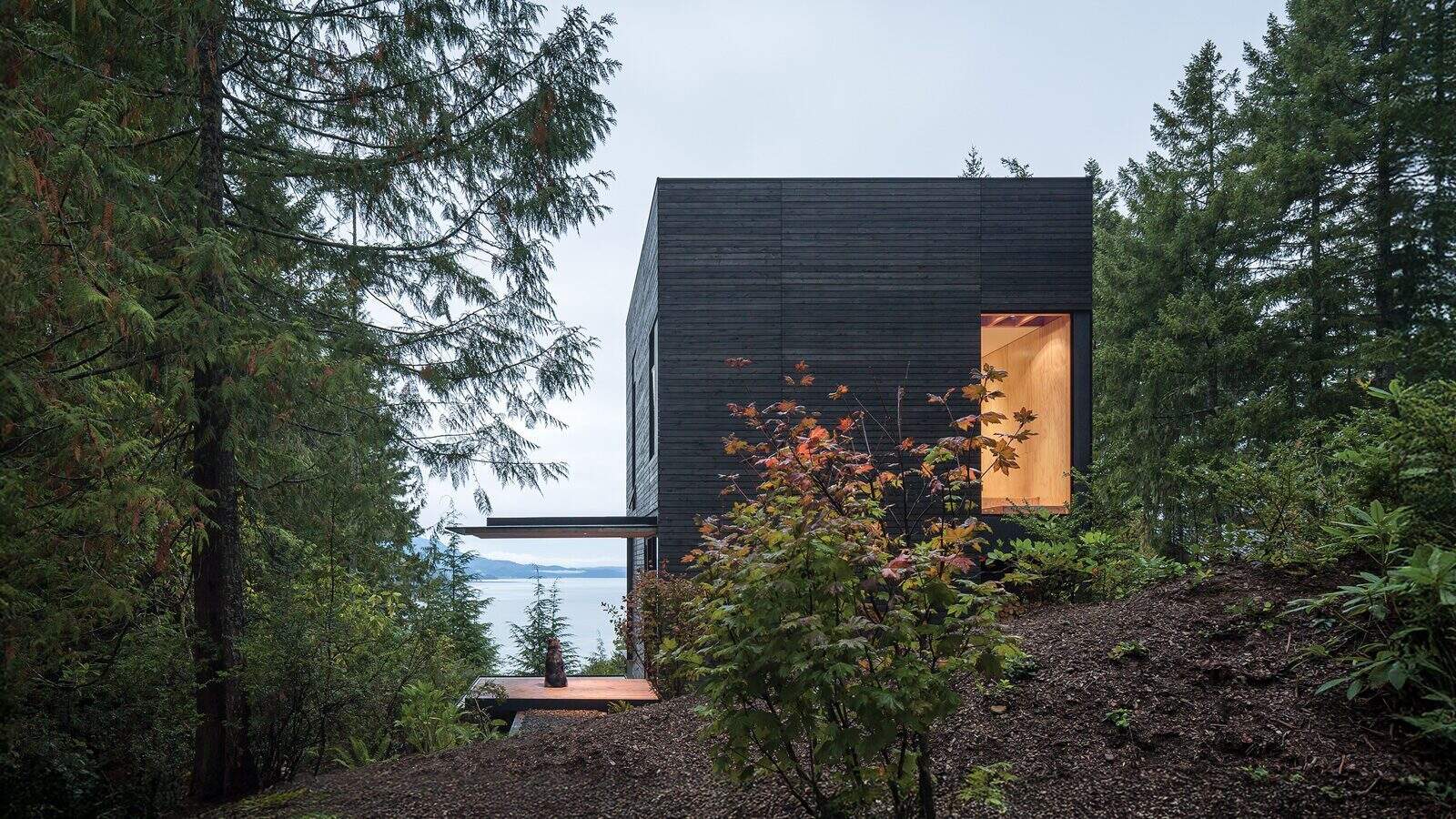 LittleHouse_mwworks-004_1600x900 A modern, dark wood cabin with large windows sits on a forested hillside—ideal for second homes or summers in the waterfront—partially obscured by trees and foliage, overlooking the water in the background.
