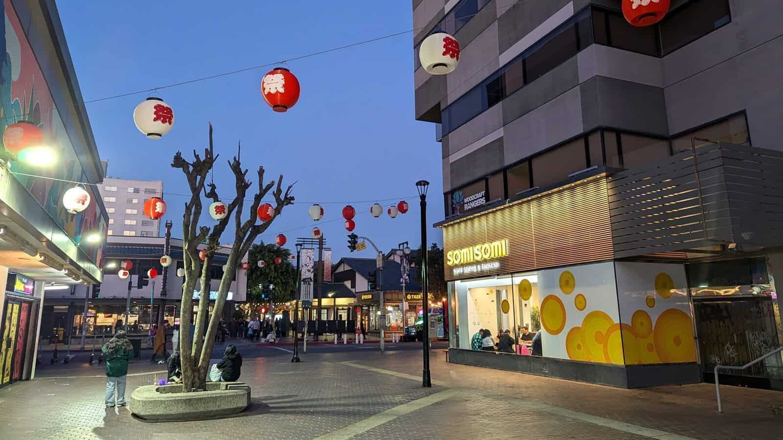 Outdoor plaza at dusk with Japanese lanterns overhead, people sitting on benches, and the SomiSomi dessert shop on the right, where Hope Rises in L.A. as smoke fades into the evening sky.