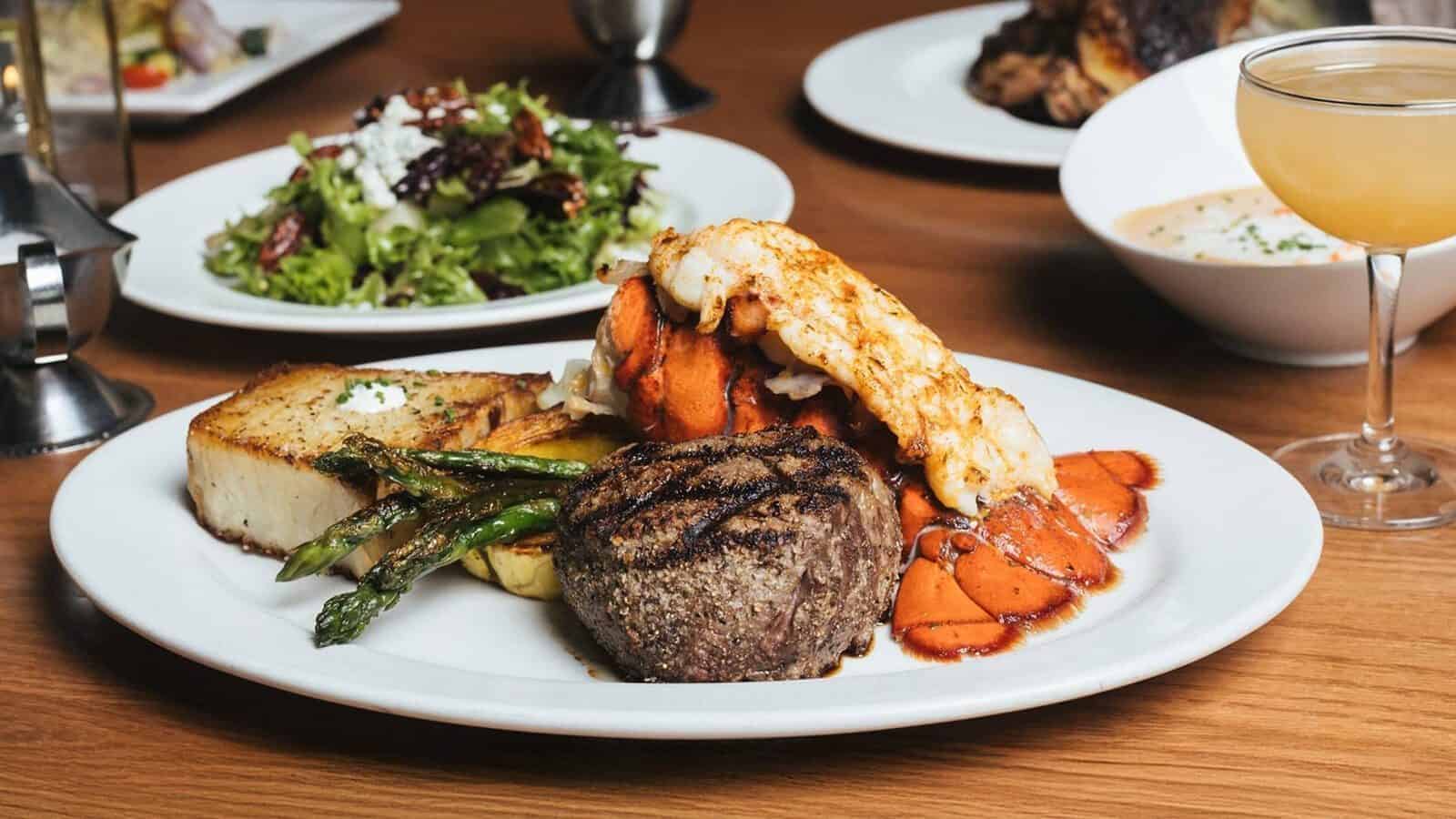 A plate with grilled steak, lobster tail, asparagus, roasted potatoes, and lemon—perfect for savoring during Heart of Glass: A Weekend in Tacoma—with salad, soup, and a cocktail on the side.