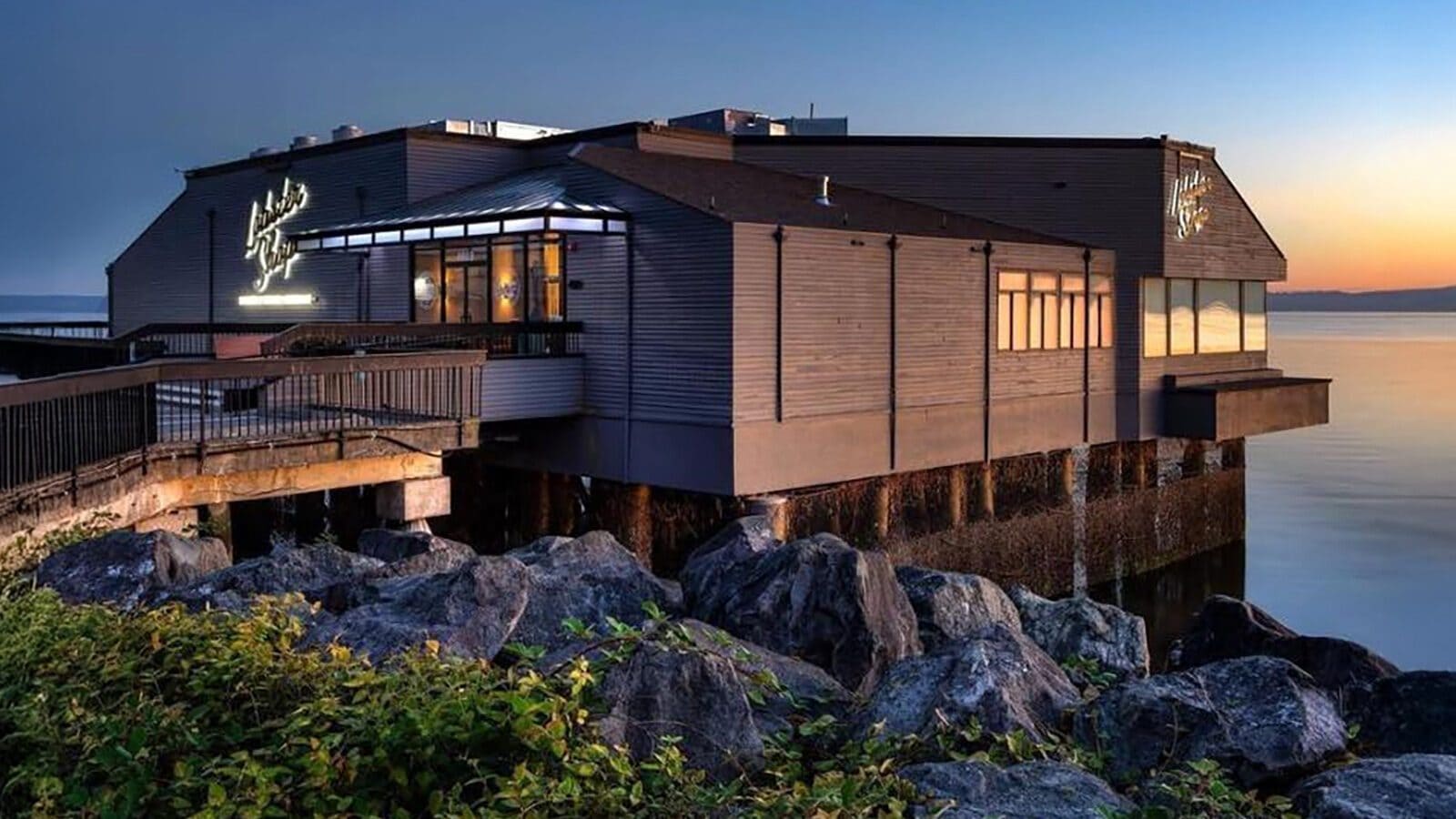 A waterfront restaurant building extends over rocks and water at sunset, its neon sign glowing like Heart of Glass: A Weekend in Tacoma, with large windows facing the shimmering waves.