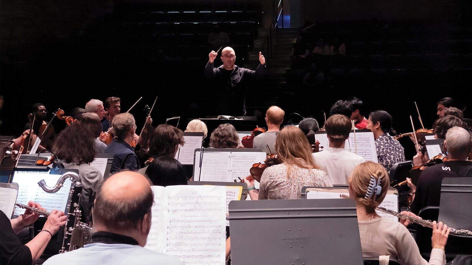 Mahler-Fest_1600x900 A conductor leads an orchestra during a rehearsal at the Shorewood Performing Arts Center, with musicians reading sheet music beneath dim lights as they prepare for the Mahler Fest 30th anniversary.