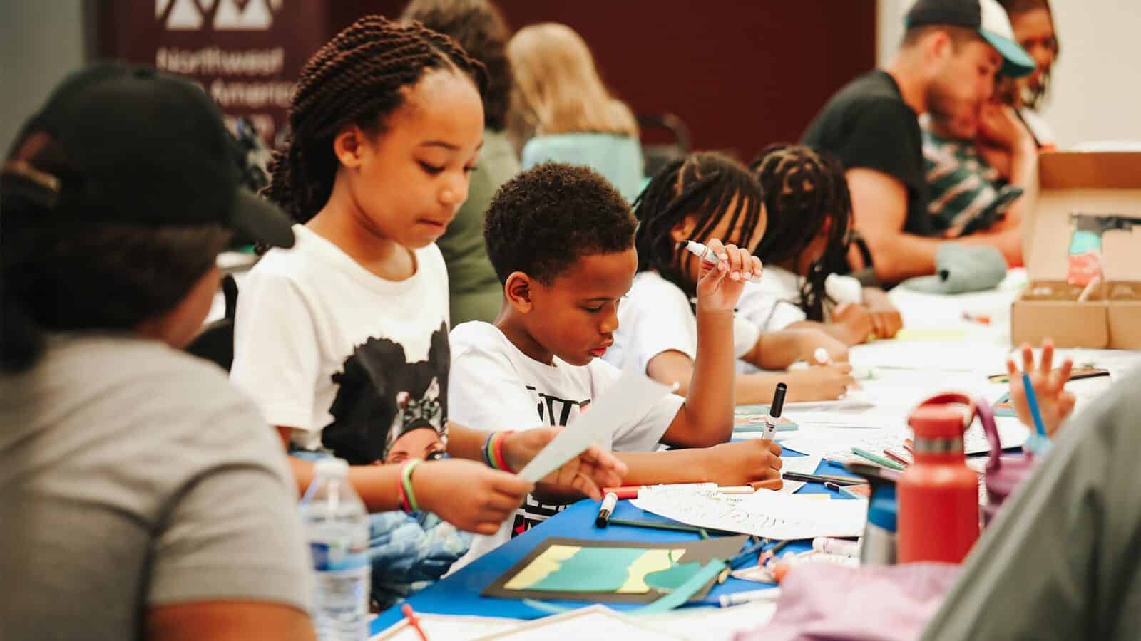 Children sit at tables working on arts and crafts projects with paper, markers, and other supplies—a fun classroom activity for anyone seeking ideas for what to do in Seattle with kids.