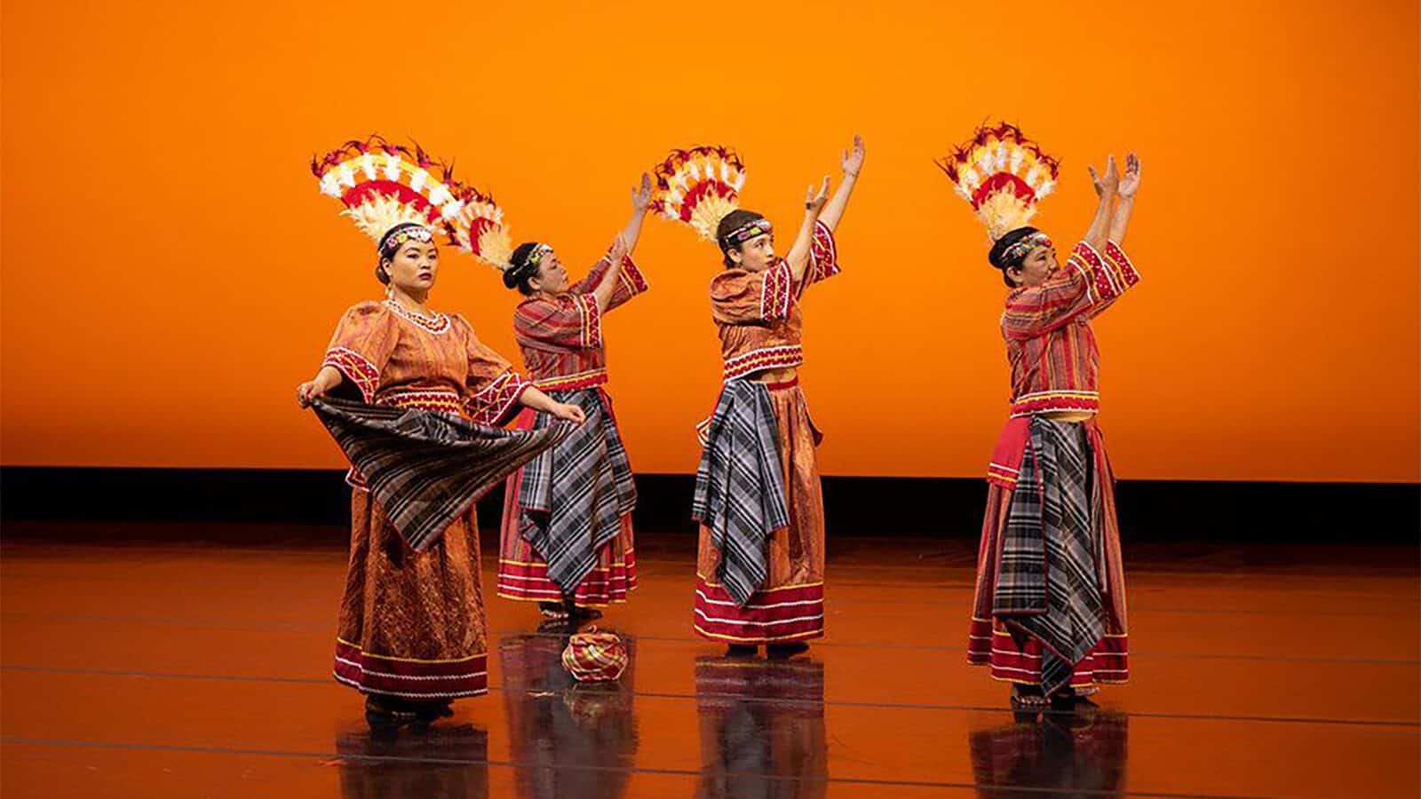 Set your sights on these summer festivals across Puget Sound, where four dancers in traditional attire with feathered headpieces perform on stage against an orange backdrop, raising their arms in unison.