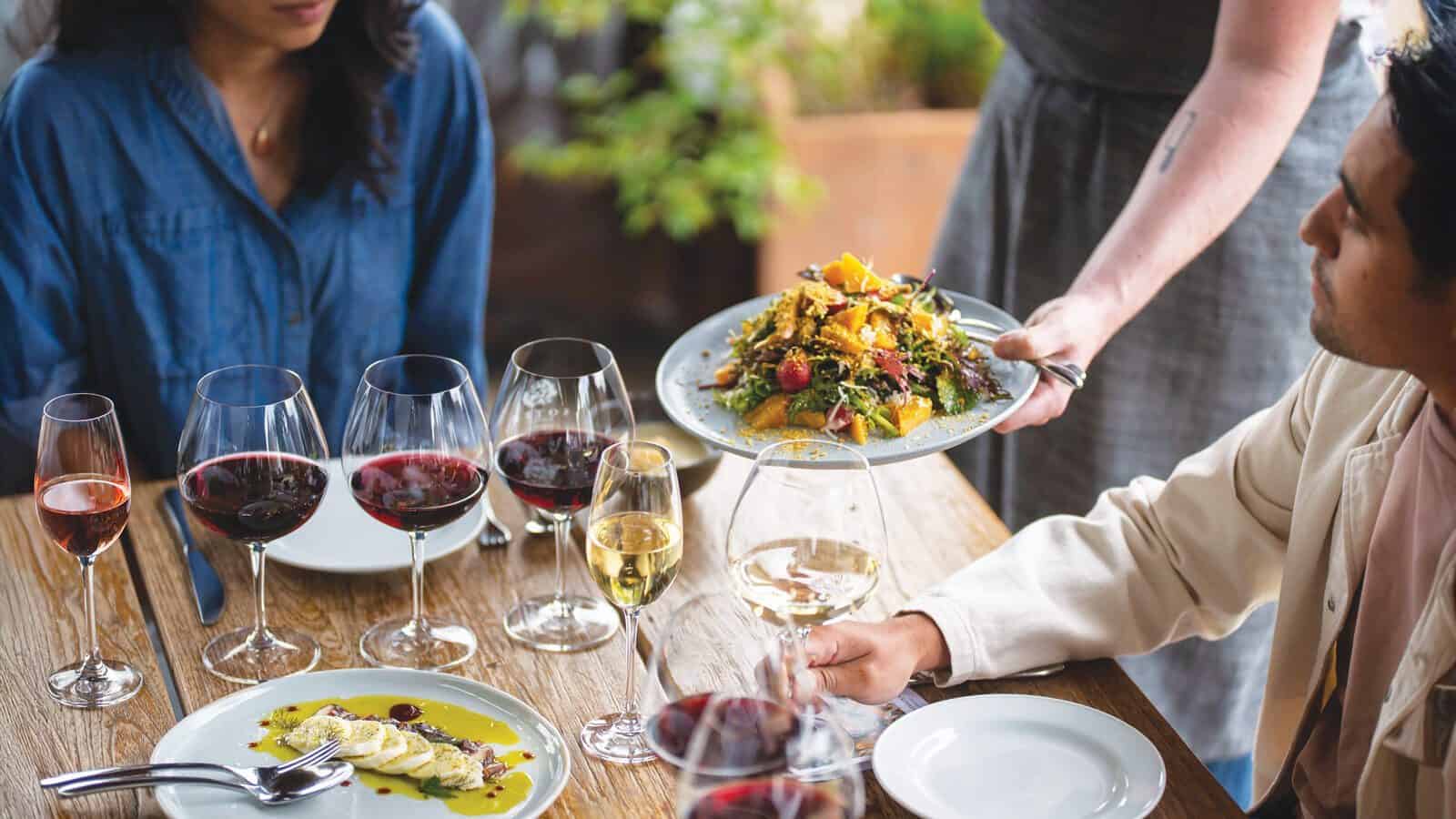 A person serves a plate of salad to a man at a table set with wine glasses, plates, and another seated woman, evoking the relaxed elegance of Oregon wine country dining.