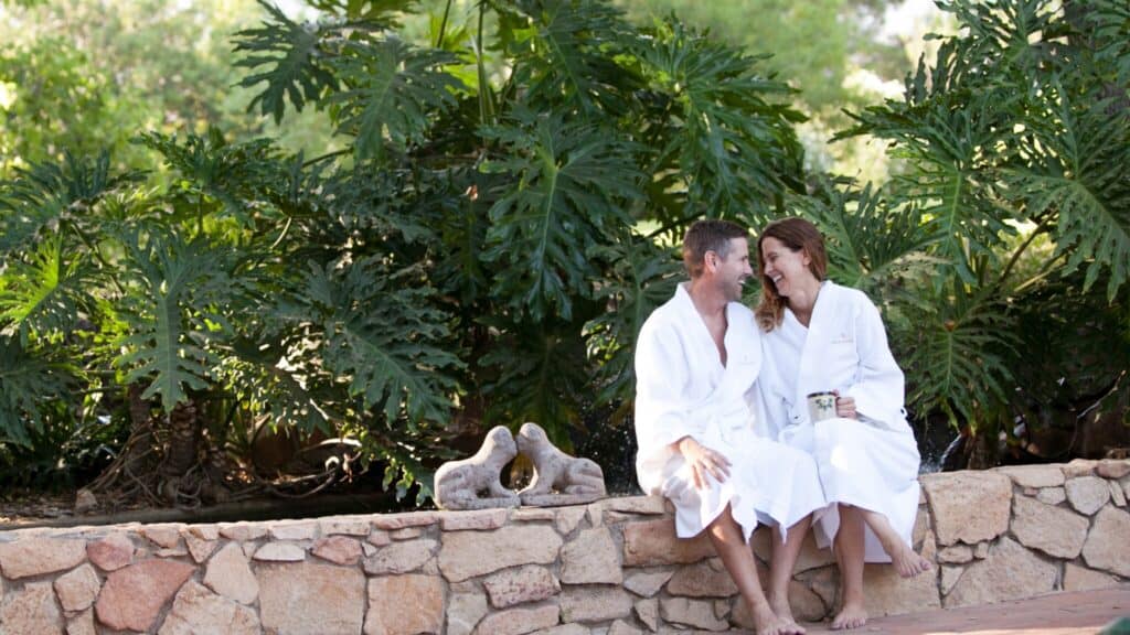 A man and woman wearing white robes sit on a stone wall outdoors at Rancho La Puerta, surrounded by lush green plants. They are smiling, leaning towards each other in this tranquil sanctuary of holistic health.