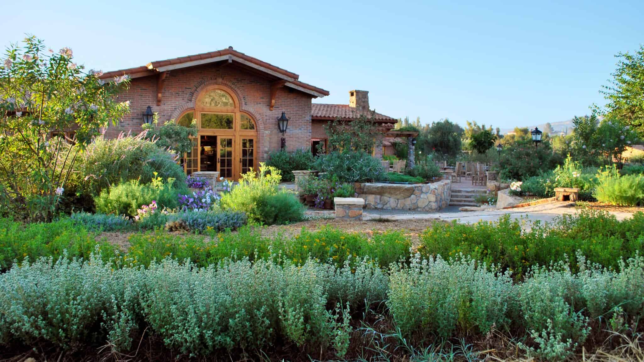 RLP_2019_cocina (edited) A rustic house with an arched window is surrounded by lush gardens, shrubs, and stone pathways under a clear blue sky, creating a sanctuary reminiscent of Rancho La Puerta.