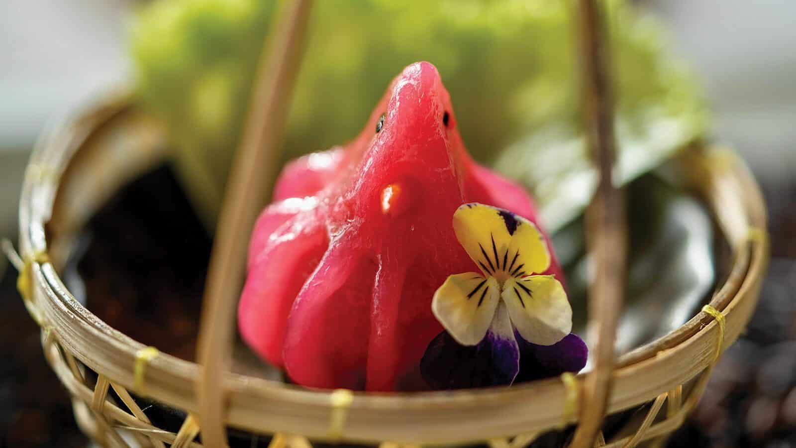 A pink, bird-shaped dessert with a flower garnish sits in a woven basket with green leaves in the background—a memorable meal perfect for a culinary getaway in beautiful British Columbia.