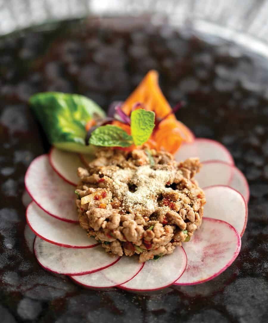 Minced meat salad garnished with mint and vegetables, served atop thinly sliced radishes in a circular pattern on a dark plate—a memorable meal inspired by the culinary getaway experiences of British Columbia.
