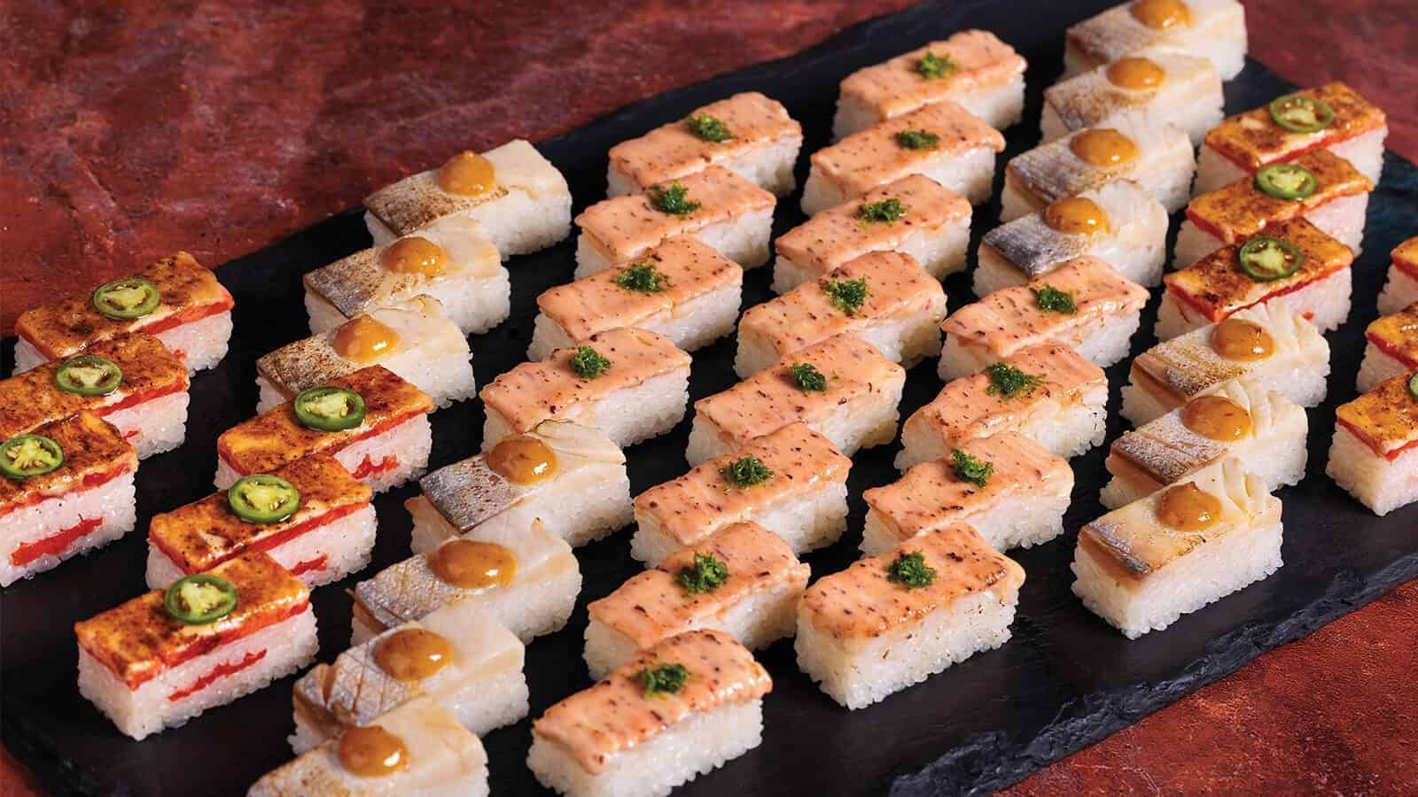 Rows of rectangular sushi pieces with unique toppings—sauces, herbs, jalapeño slices, and grilled fish—are arranged neatly on a black serving tray, perfect for a memorable meal during your culinary getaway.