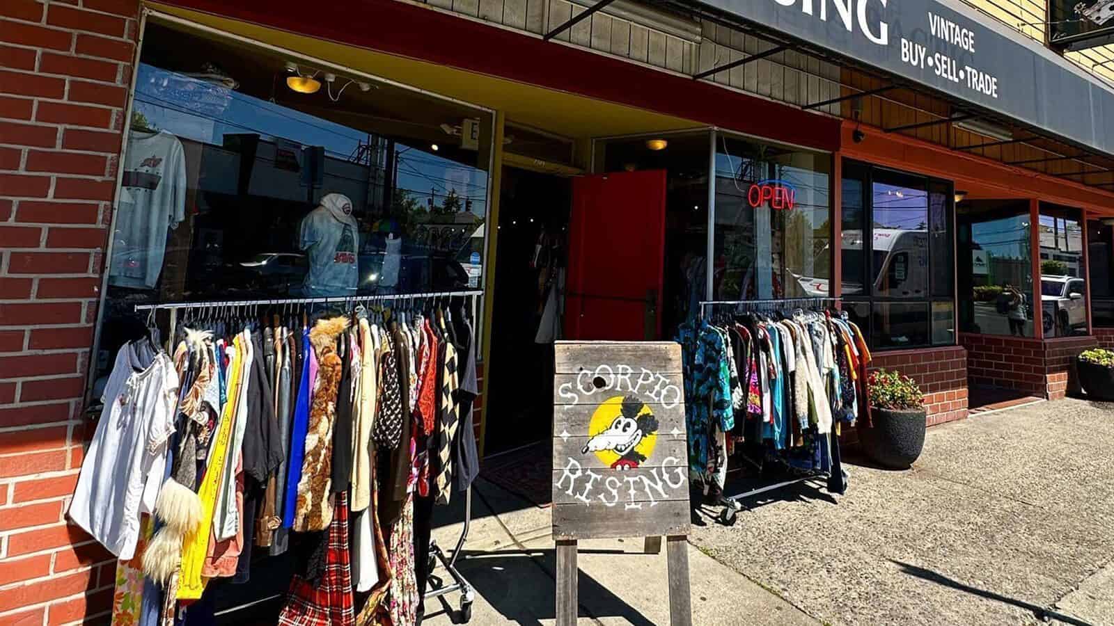 Exterior of a vintage clothing store in "Heart of Glass: A Weekend in Tacoma," with racks of colorful clothes outside, a chalkboard sign reading "CrowPot RISING," and an open door inviting customers in.
