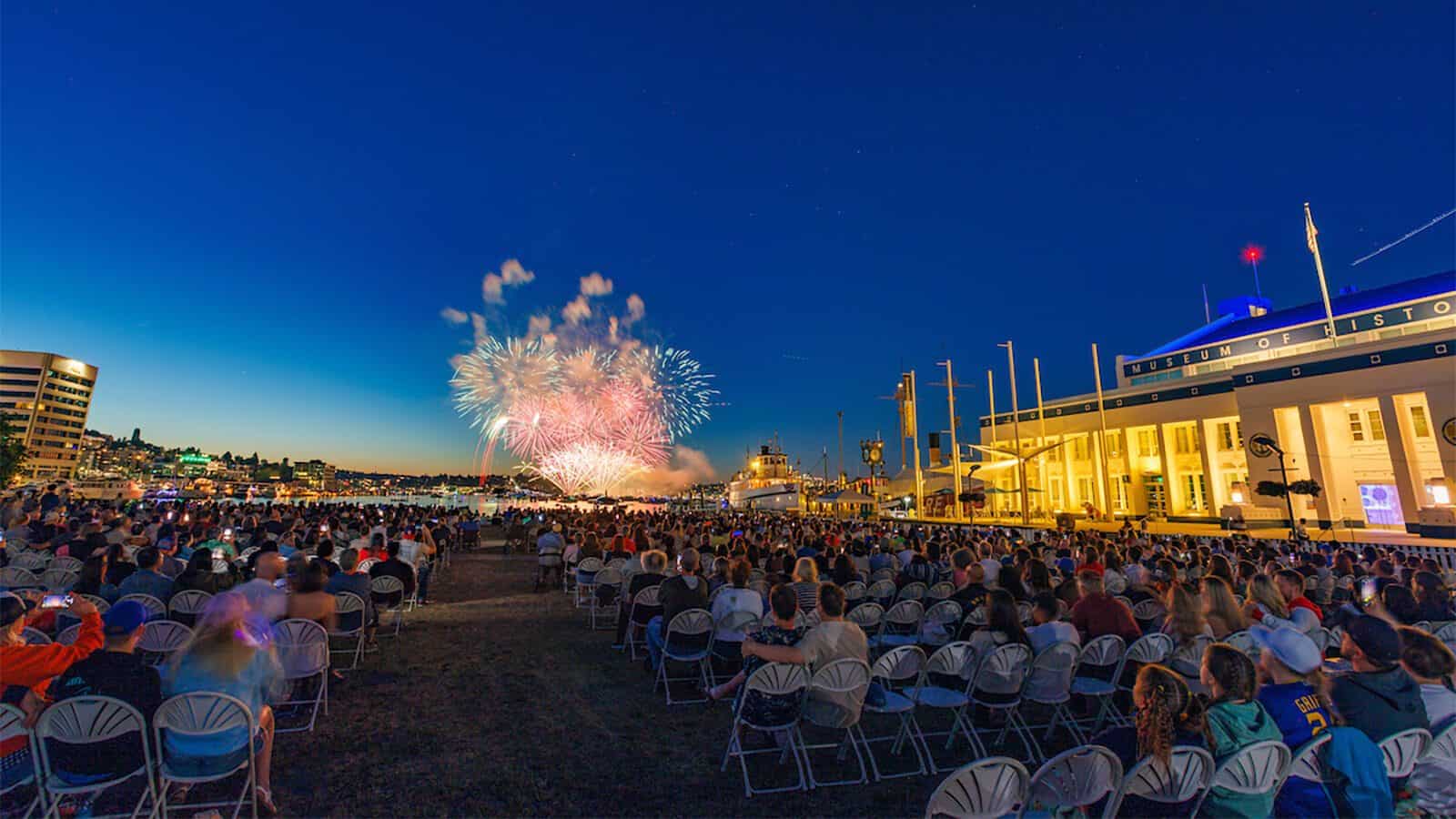 Set your sights on these summer festivals across Puget Sound, where a crowd seated outdoors at dusk watches fireworks burst over the waterfront near a large white building and docked boats.