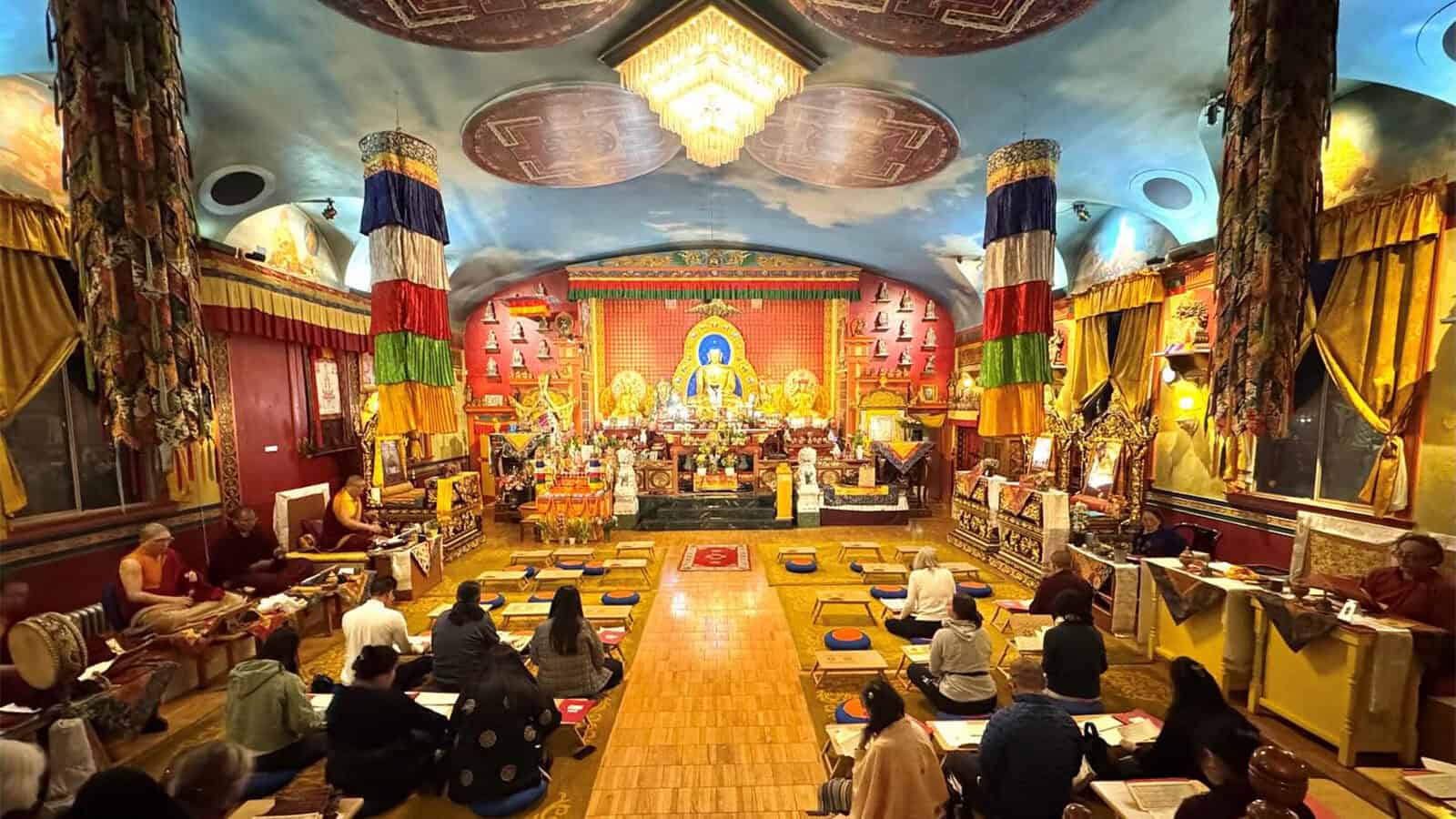A group of people sit on cushions in a richly decorated temple, facing an altar with a large Buddha statue and colorful banners—a serene scene reflecting Seattle heritage and its vibrant AANHPI community.