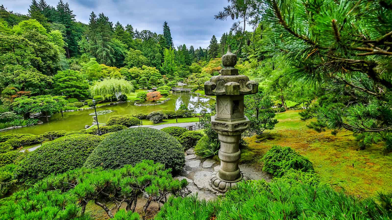 A Japanese garden featuring a stone lantern, manicured shrubs, a pond, and dense green trees under a cloudy sky—one of Seattle cultural landmarks celebrating the AANHPI community’s heritage and tranquility.
