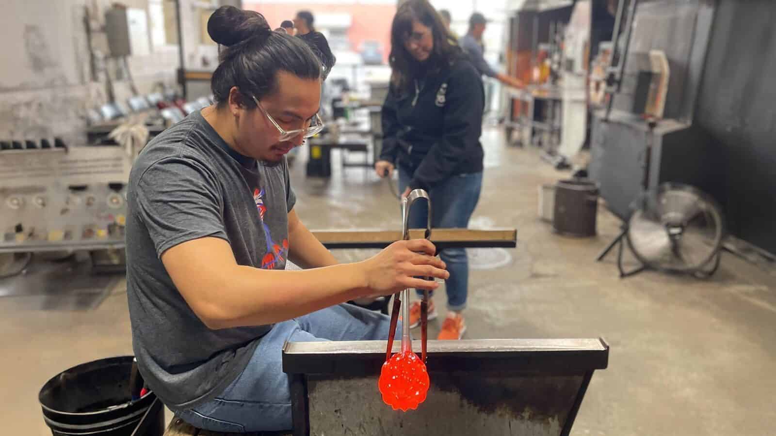 A person shapes hot glass with tongs at a workshop table, capturing the spirit of Heart of Glass: A Weekend in Tacoma, while another stands in the background in an industrial studio setting.