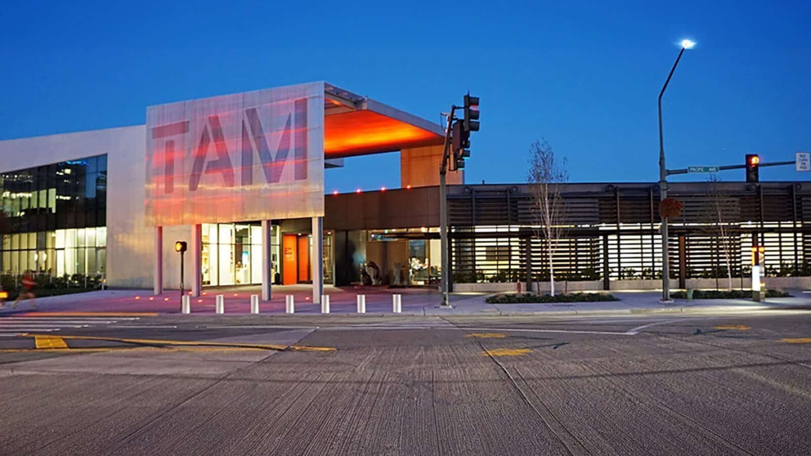 A modern building with "TAM" signage at dusk, featuring large windows and a lit entrance—capturing the vibrant spirit of Heart of Glass: A Weekend in Tacoma—situated at a bustling street intersection in the foreground.