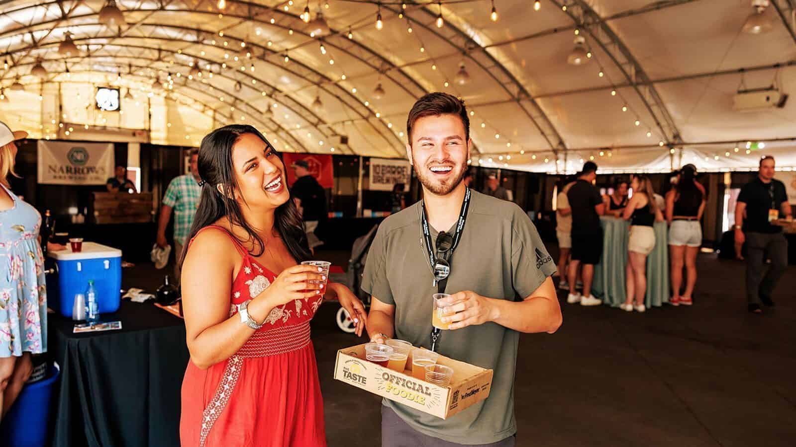 Set your sights on these summer festivals across Puget Sound, where a woman in a red dress and a man with a badge and tray of sample cups smile and hold drinks inside a festive, string-lit event tent.