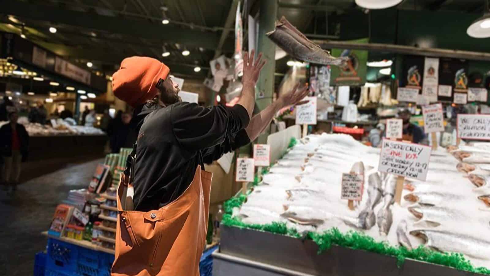 Tourism-Image-PikePlaceMarket_1600x900 A fishmonger wearing an orange hat and apron throws a fish in the air at an indoor seafood market, celebrating the lively atmosphere as The Numbers Are In: Seattle Tourism Is Back, with various fish displayed on ice.