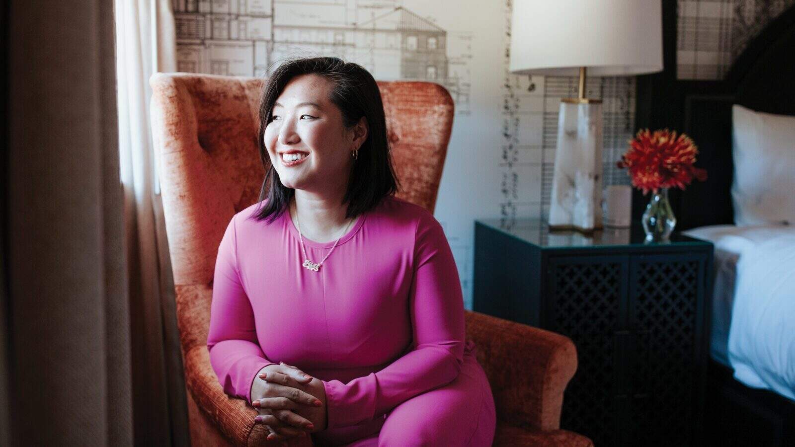 Trailblazing Women: Stephiney Foley sits in a bright pink outfit on an orange chair, smiling as she looks toward the window. A lamp and a vase with red flowers adorn the table beside her.