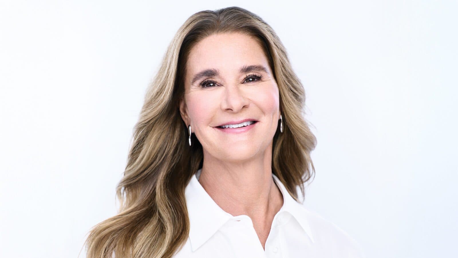 Trailblazing2025_Melinda-French-Gates_1600x900 A woman with long, wavy brown hair and light skin smiles at the camera. She is wearing a white blouse and small drop earrings, embodying the grace often associated with Melinda French Gates of the Gates Foundation, against a plain white background.