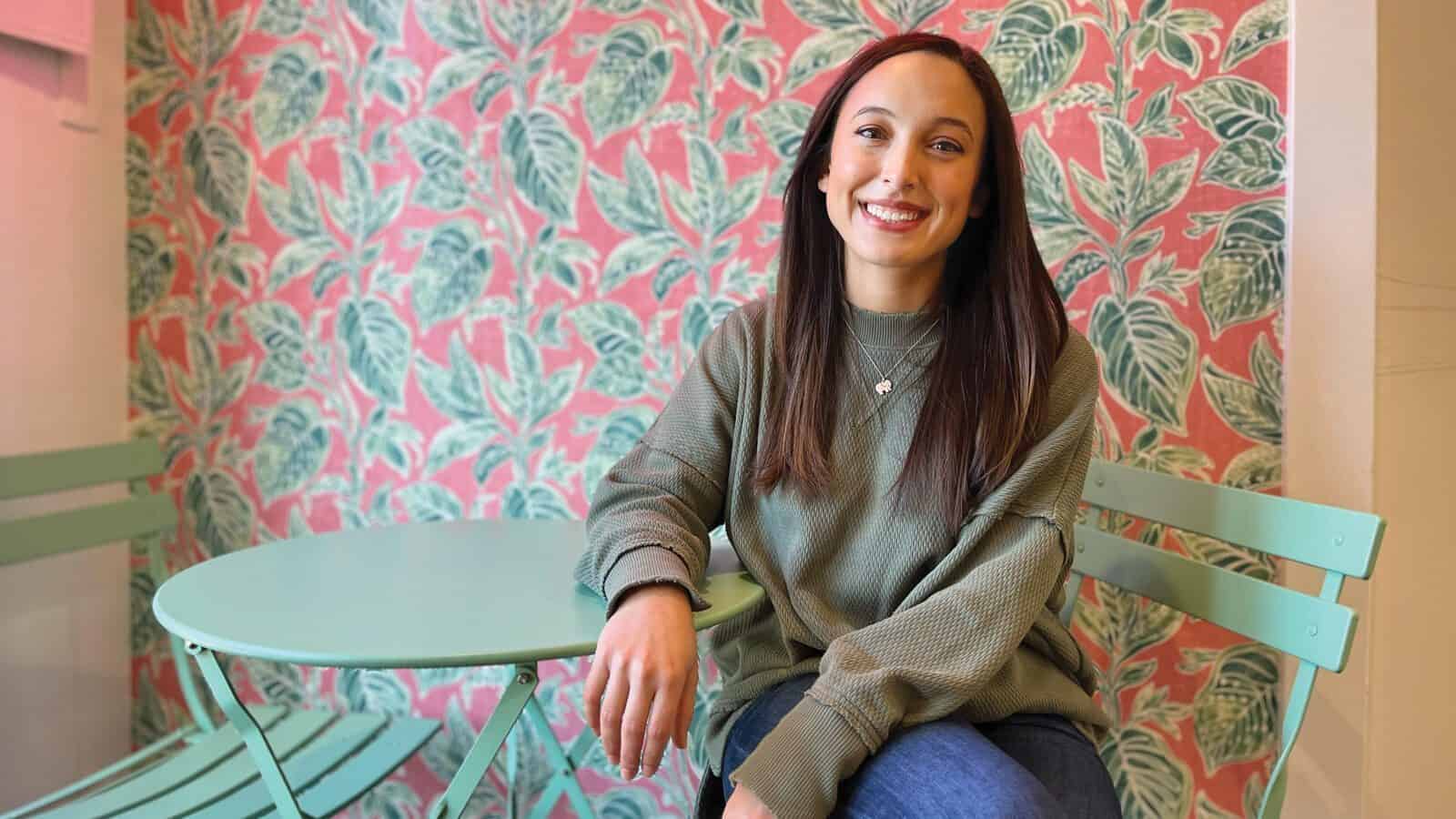 Trailblazing2025_Rachel-Barnecut_1600x900 Rachel Barnecut sits at a mint green table and chair set in front of pink and green leaf-patterned wallpaper, smiling at the camera—a fitting portrait among trailblazing women.