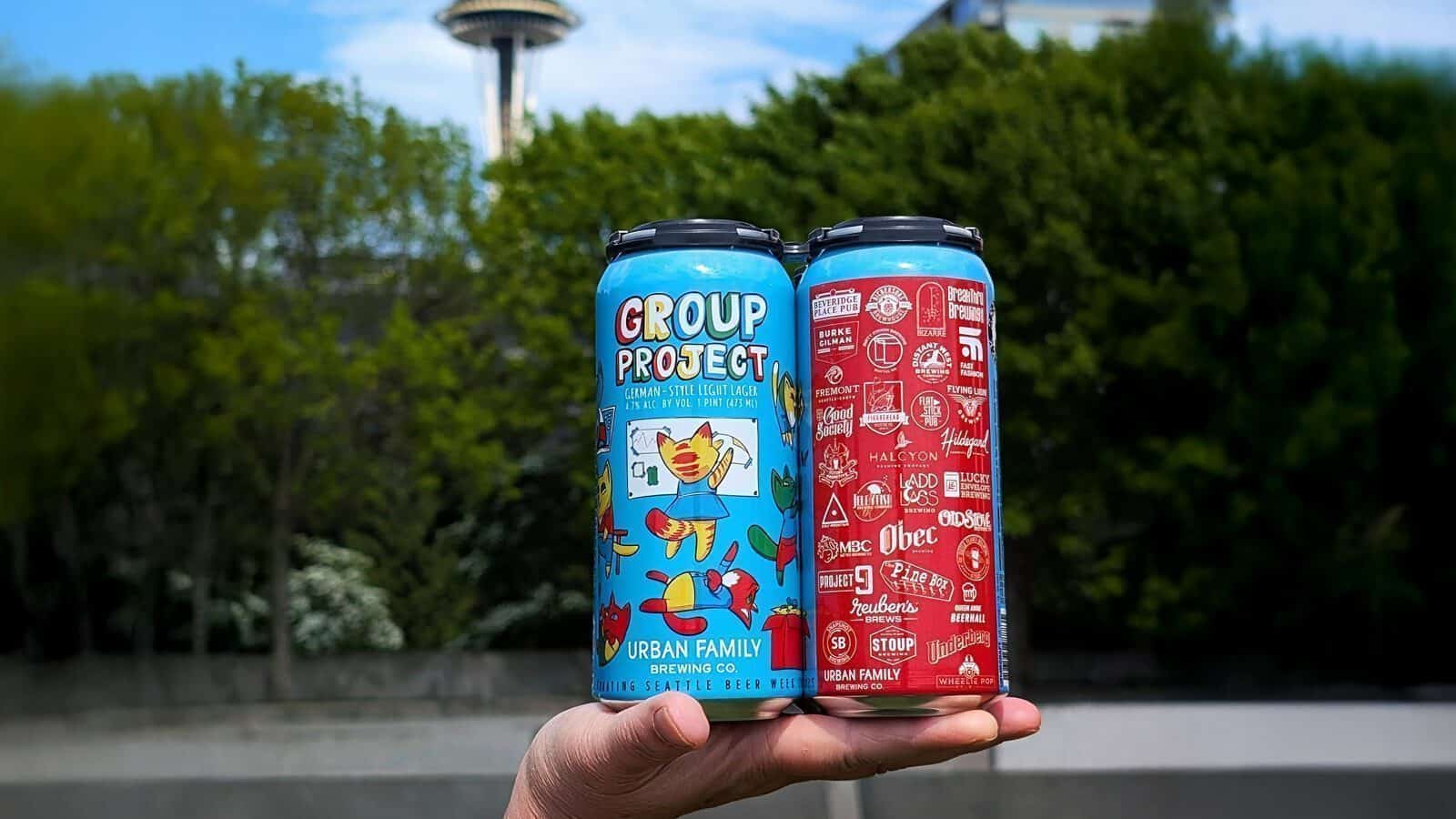 UrbanFamilyBrewingCo-BeerCans_1600x900 A hand holds two colorful cans of Urban Family Brewing beer outdoors, with the Space Needle and green trees in the background—perfect for celebrating Seattle Beer Week.