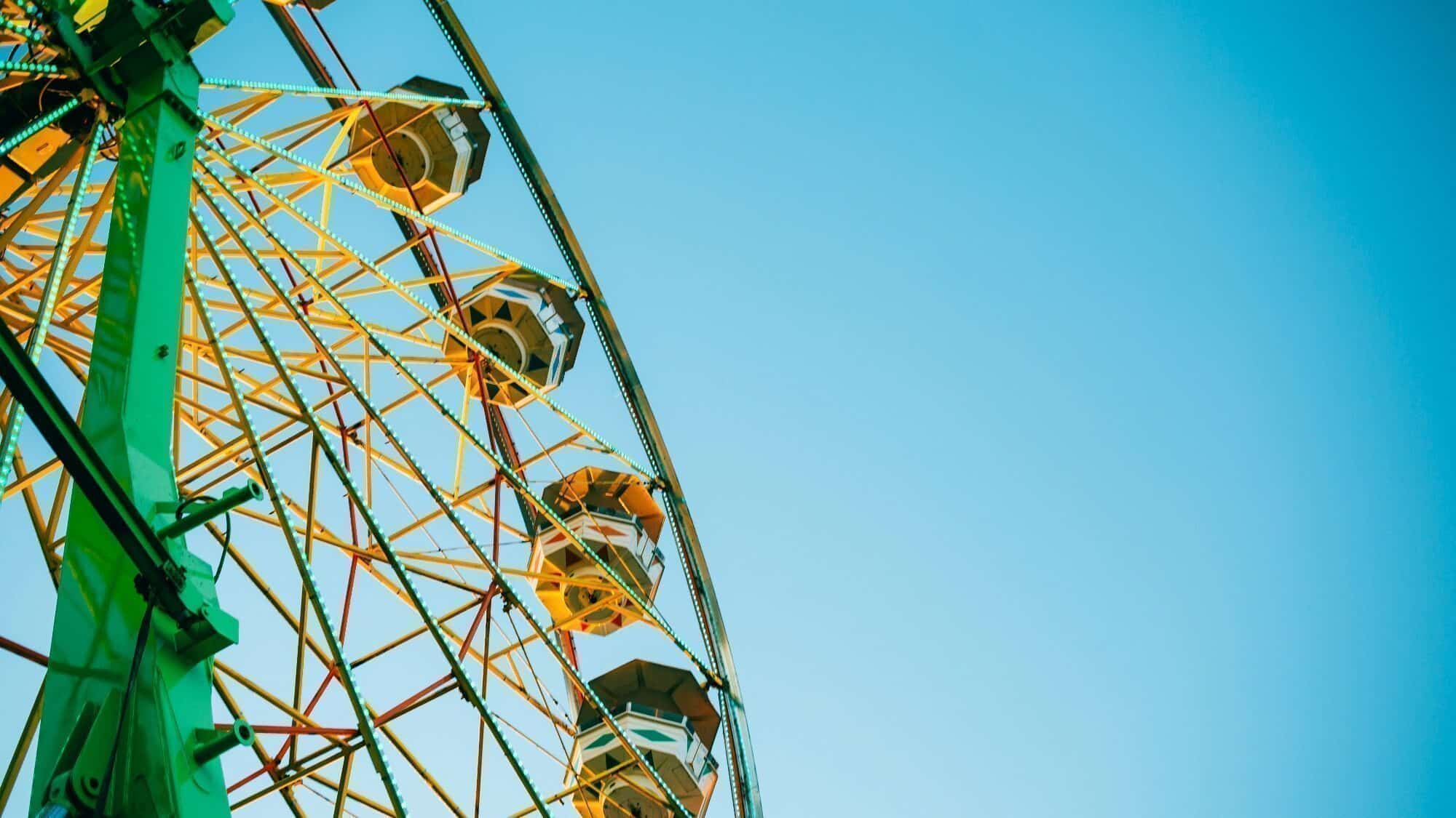 Partial view of a Ferris wheel with yellow and white metal framework and several gondolas against a clear blue sky.