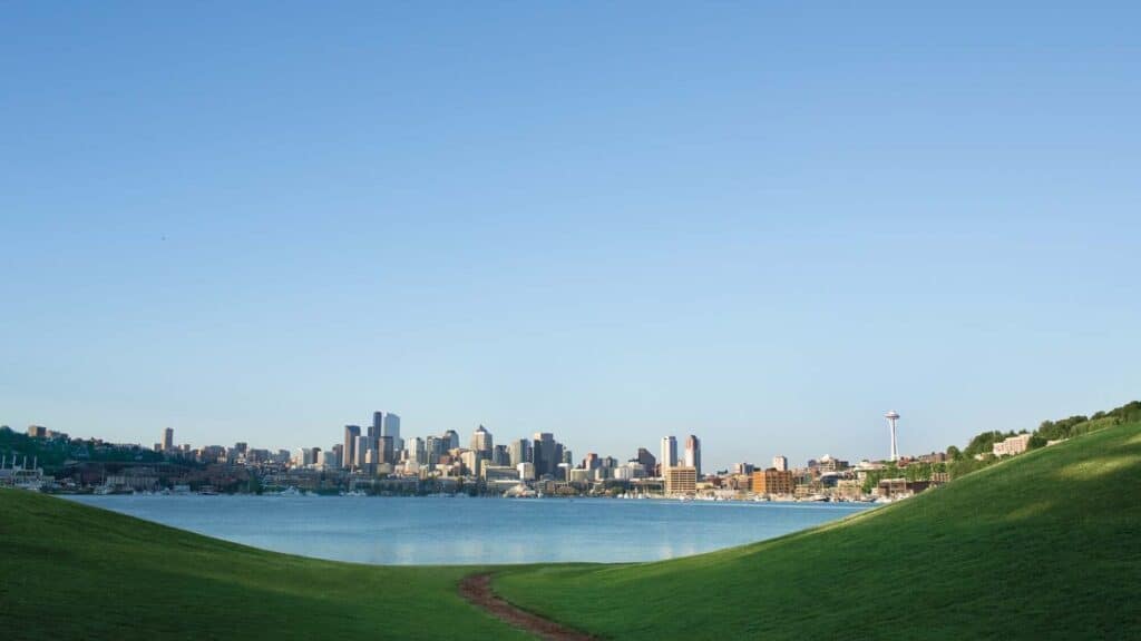 A view of a city skyline across a body of water, framed by two grassy hills under a clear blue sky.