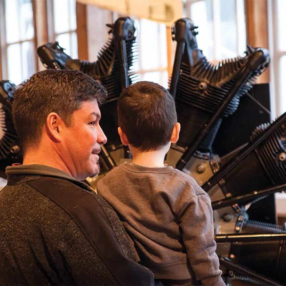 An adult and a child stand in front of a large, radial aircraft engine display indoors, both facing away from the camera—a Must List experience to remember on 06-12-2025.
