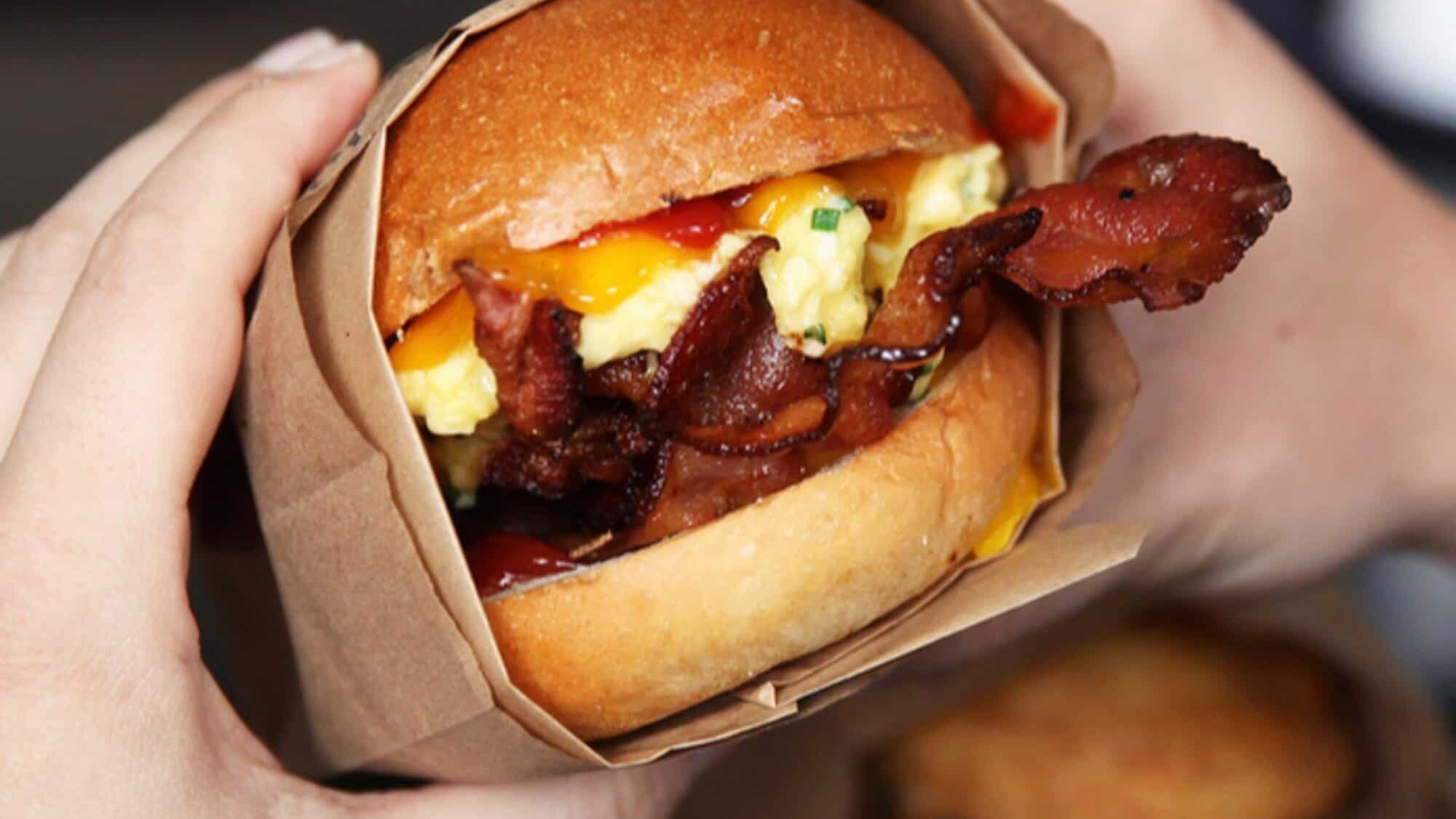 EGGSLUT Quality+&+Taste-square-2000x2000-16-9-2000x1125 A person holds a sandwich with crispy bacon, scrambled eggs, melted cheese, and sauce in a brown paper wrapper.