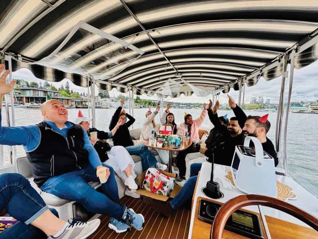 A group of people wearing party hats sits around a table on a covered boat during Seattle summer, smiling and raising their hands, with water and city buildings visible in the background.