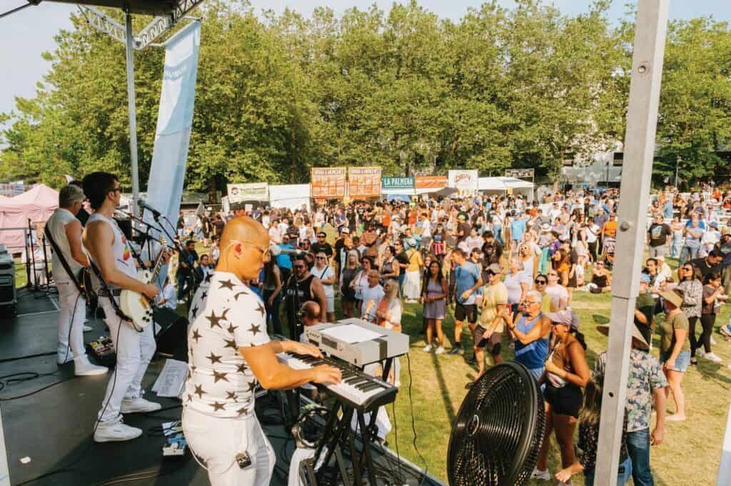 A band performs on an outdoor stage to a lively crowd at the Bite of Seattle festival, with vendor booths and trees visible in the background.