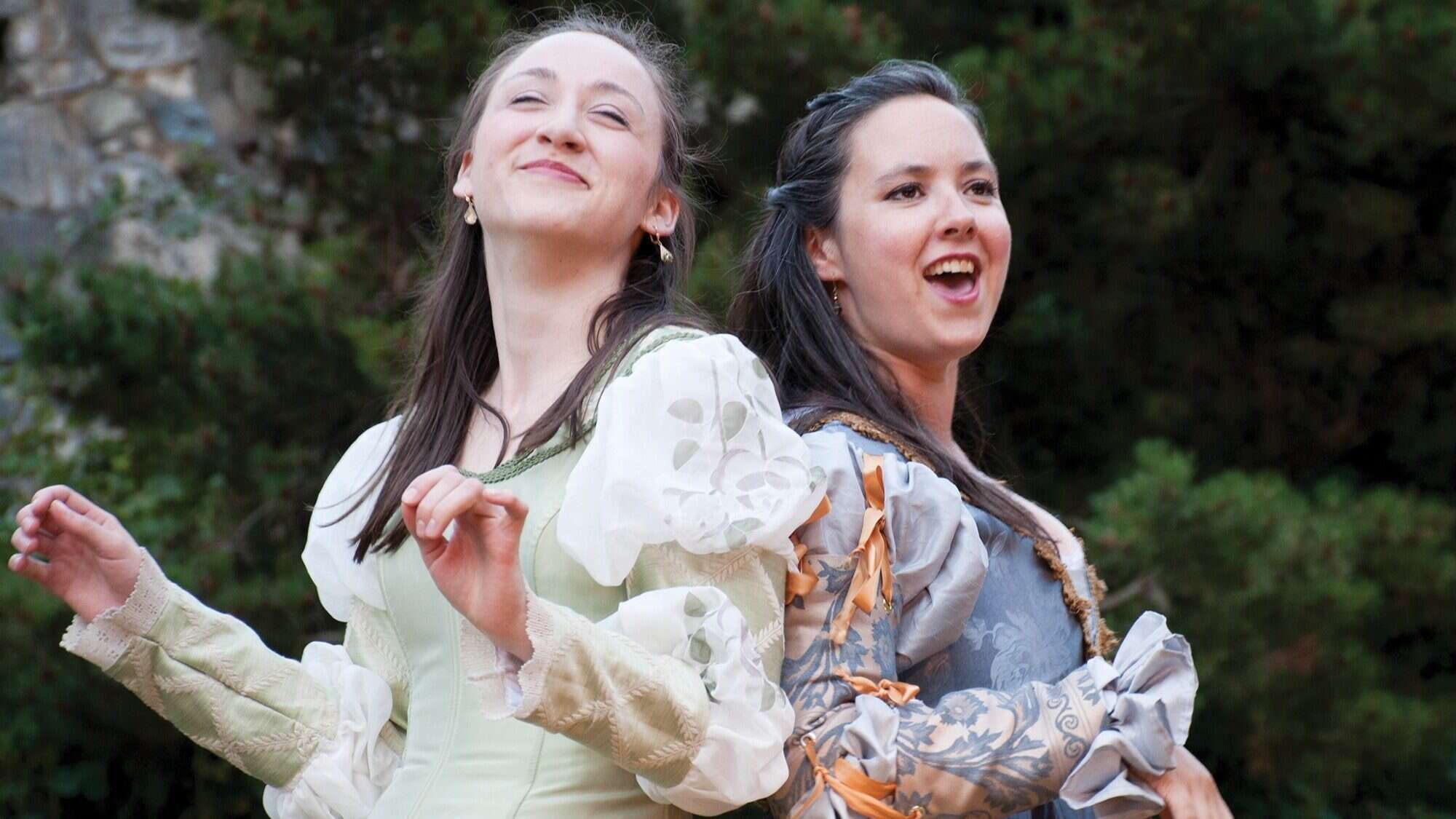 ESSENTIALS_Shakespeare-16-9-2000x1125-min Two women in historical costumes stand back to back outdoors, smiling and gesturing with their hands at a lively food festival, surrounded by greenery in the background.
