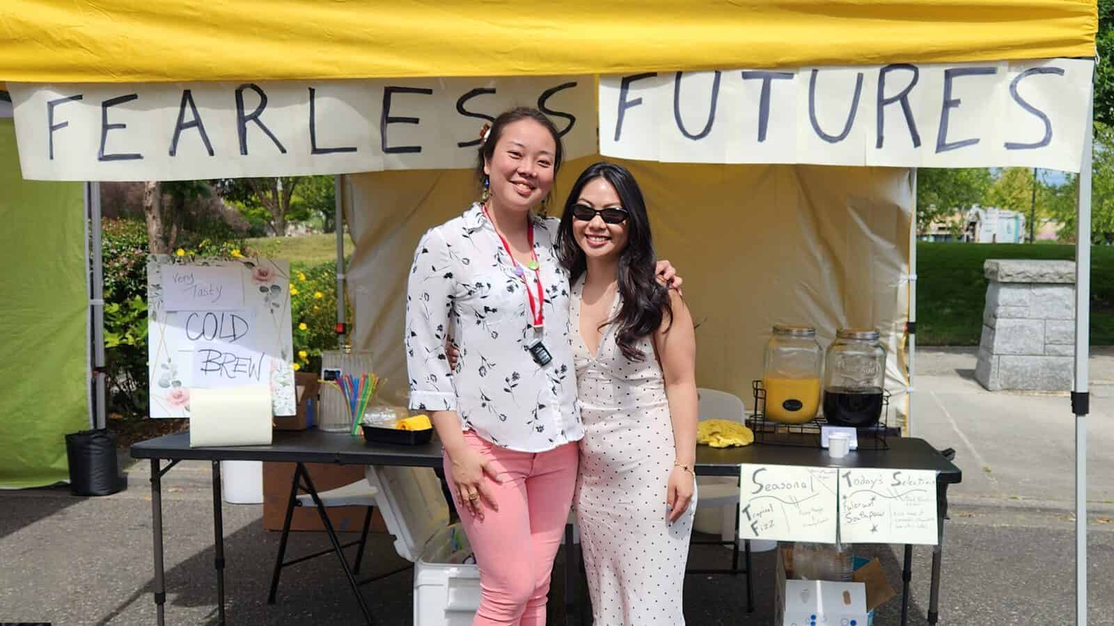 FearlessFuture-LaunchDay_1600x900 Two women stand smiling in front of a coffee stand with a "Fearless Futures" banner, cold drinks, and a handwritten menu at an outdoor event, welcoming everyone.