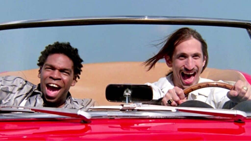 Two people laugh and smile while driving a red convertible car with the top down against a clear blue sky.