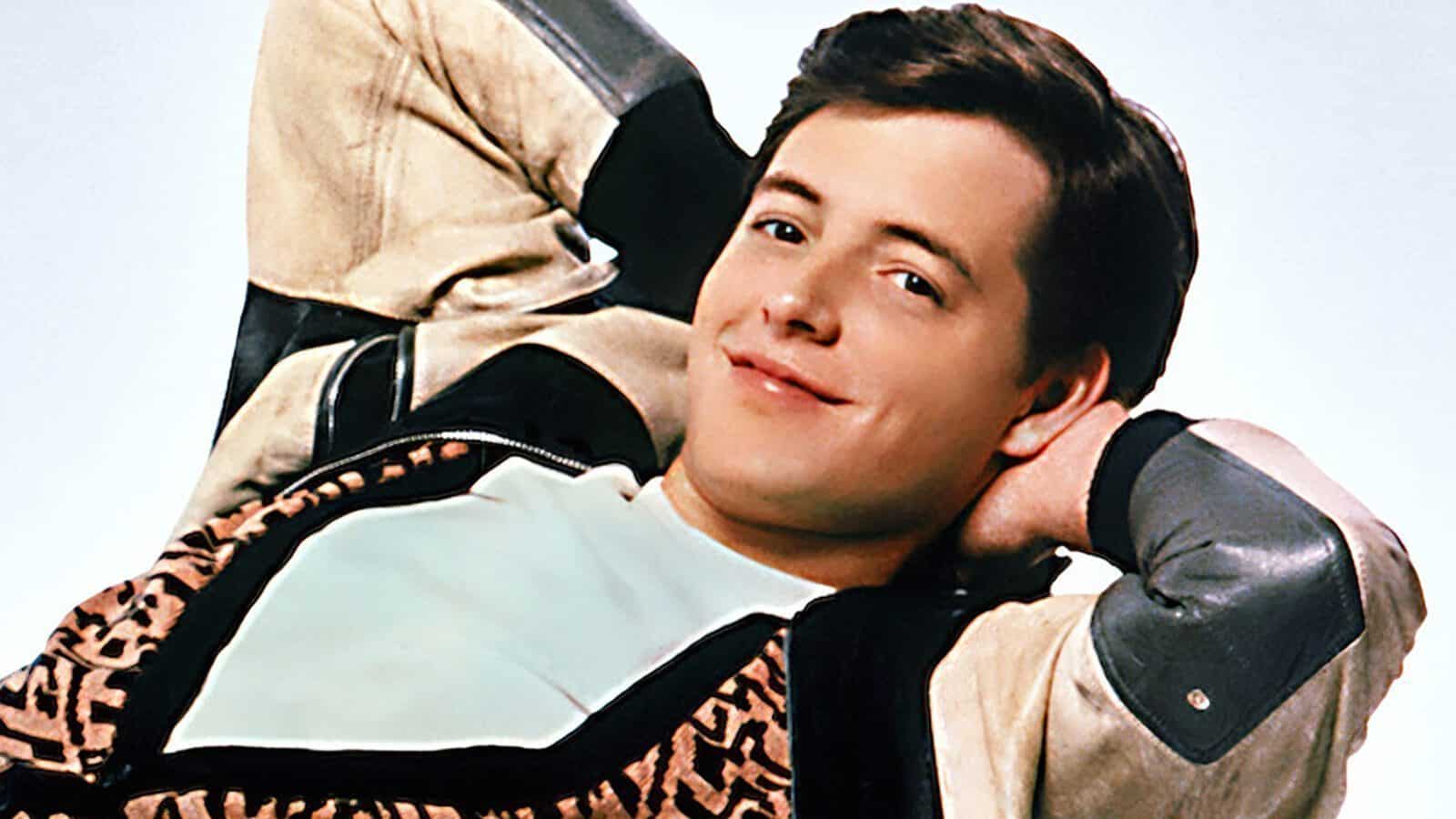 A young man with short brown hair leans back with his hands behind his head, wearing a patterned vest and a light-colored shirt and jacket, smiling at the camera.