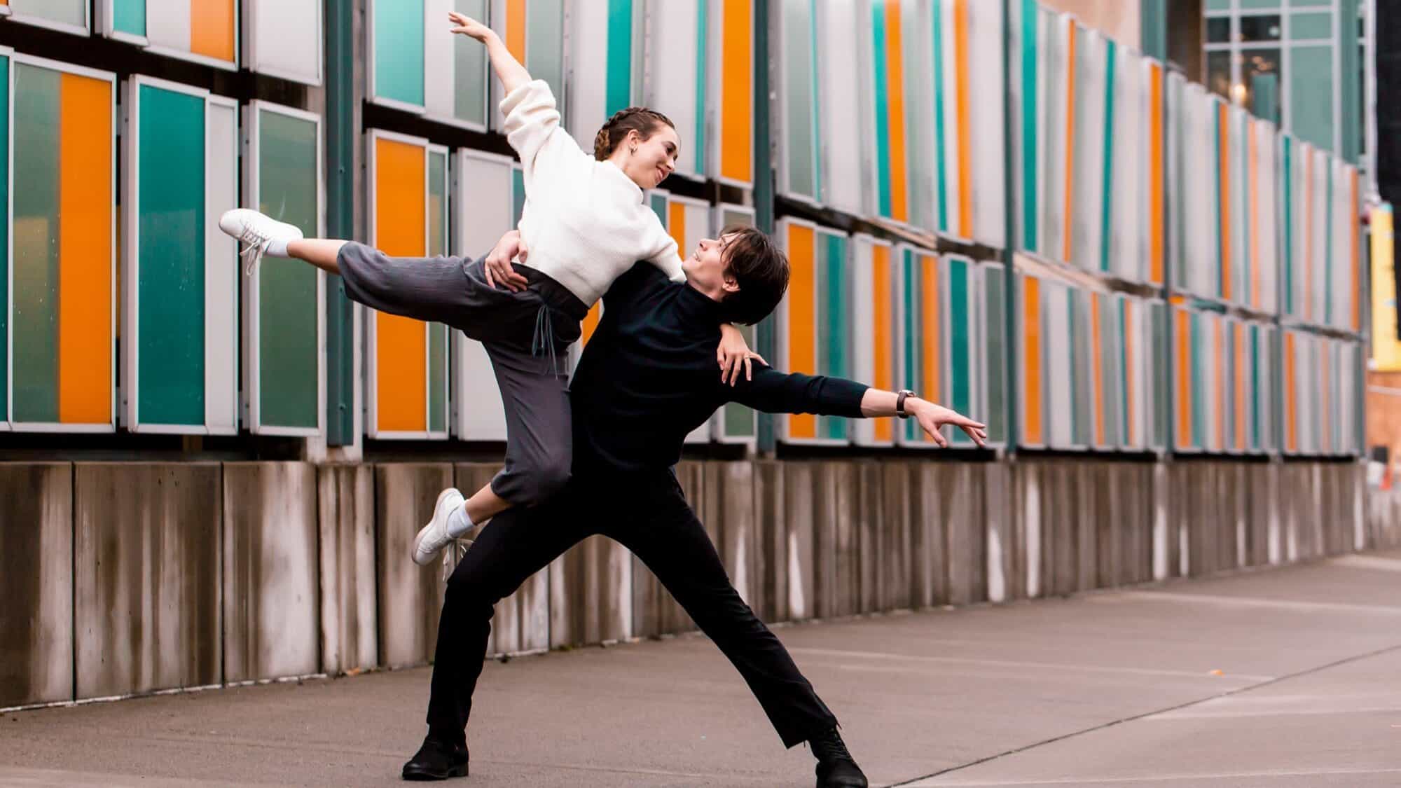 GUillame-Basso-Adage-Ballet-Ensemble-16-9-2000x1125 Two people perform a dance pose outdoors; one lifts the other, who extends an arm and leg, in front of a wall with colorful vertical panels.