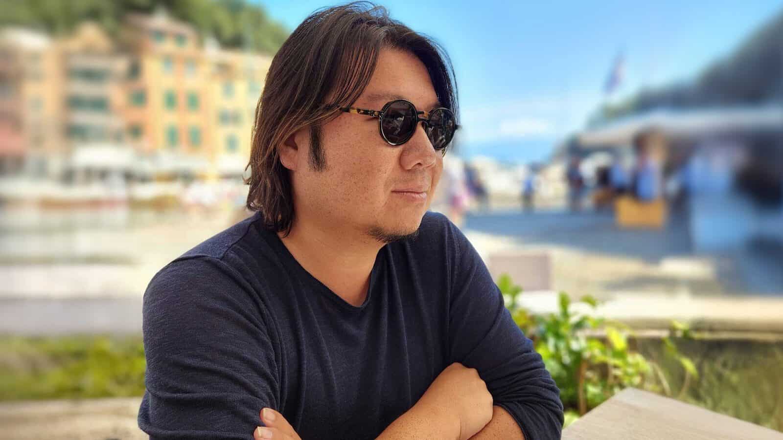 Kevin-Kwan_1600x900 A man with medium-length hair and sunglasses sits outdoors at a table with his arms crossed, evoking the stylish flair of Crazy Rich Asians, with a blurred background of buildings and people.