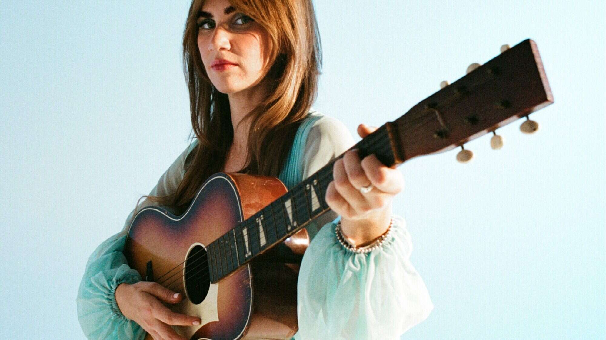 A woman with long brown hair holds an acoustic guitar against a light blue background, looking towards the camera.