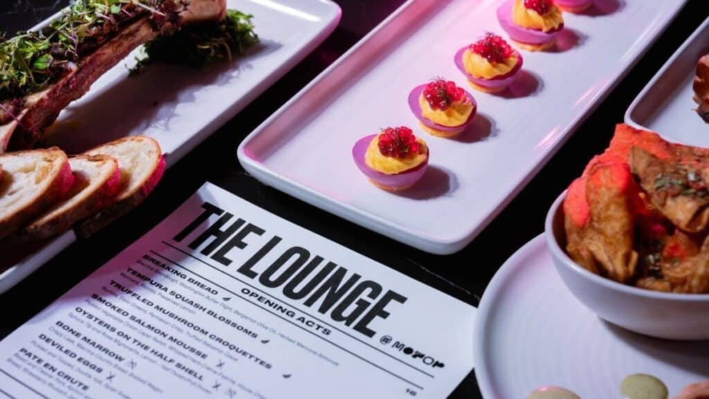 A close-up of a restaurant table with deviled eggs topped with garnish, sliced bread, chips, and a menu labeled "The Lounge Opening Acts.