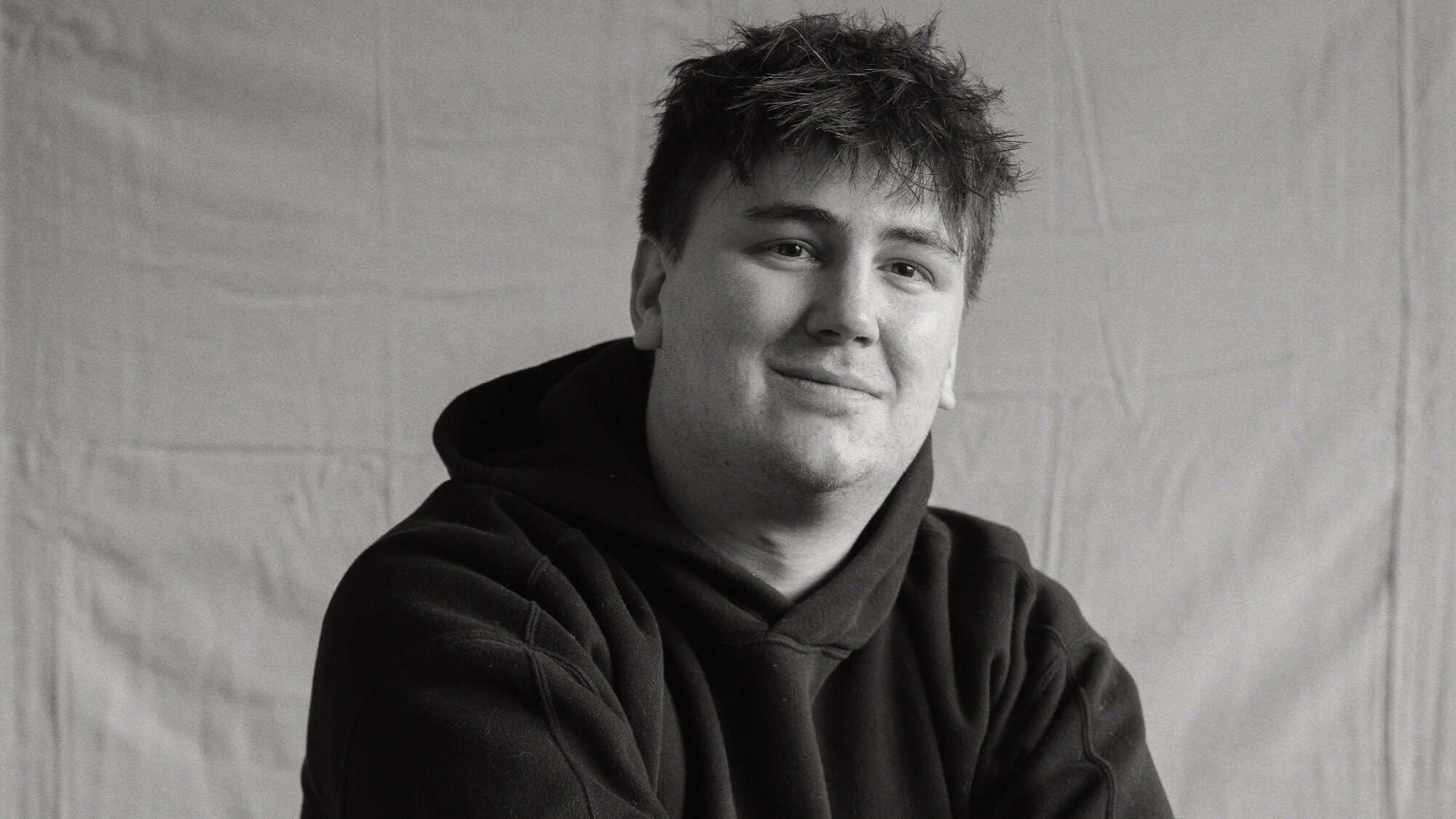 New Canlis Executive Chef James Huffman-16-9-2000x1125 A young man with short, tousled hair wearing a dark hoodie sits in front of a plain cloth backdrop, looking at the camera and smiling slightly.