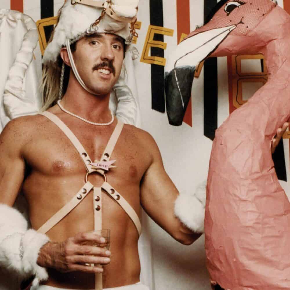 A man in a costume with harnesses and fur accents poses indoors, holding a drink next to a large pink flamingo decoration—a Must List moment you won't want to miss on 06-12-2025.