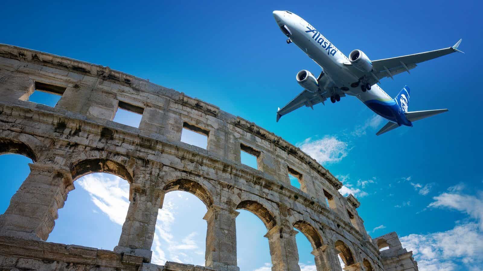 RomanColosseum-AlaskaFlight_1600x900 A commercial airplane, hinting at Alaska Airlines' new direct route to Rome starting next May, flies low over an ancient Roman amphitheater against a bright blue sky dotted with scattered clouds.