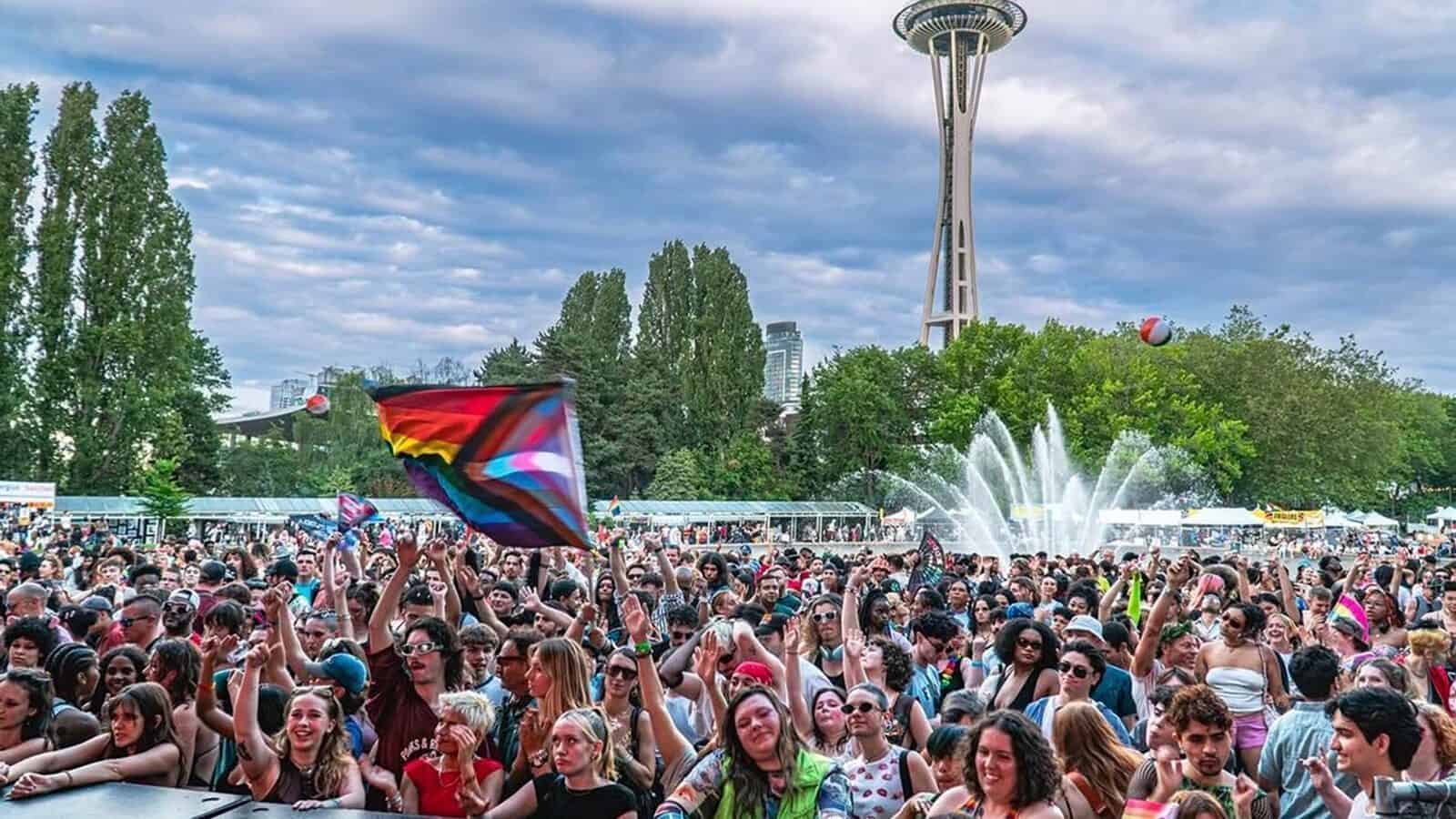 Seattle-PrideFest_1600x900 A large crowd gathers at an outdoor event near a fountain, with a Progress Pride flag visible and the Space Needle in the background on a partly cloudy day as vibrant celebrations honor Seattle’s rich LGBTQIA+ history.