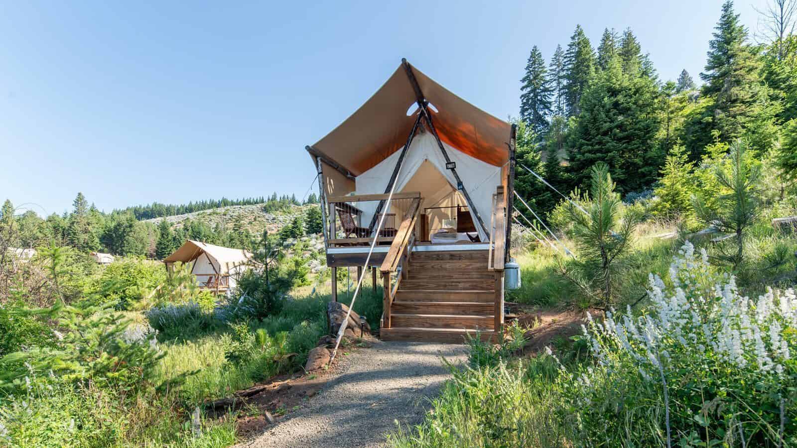 UnderCanvas-Tent_1600x900 A canvas glamping tent with a wooden deck is elevated on a platform among greenery and pine trees, creating the perfect Gorge-ous Getaway for your next forest travel adventure.